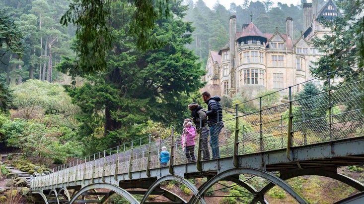 A family walking across the Iron Bridge that spans Debdon Burn between the walled garden and the house at Cragside, with a view of the Victorian mansion in the background, surrounded by trees.